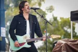 Male singer-guitarist performing outdoors, singing into a microphone while playing a mint-green electric guitar.