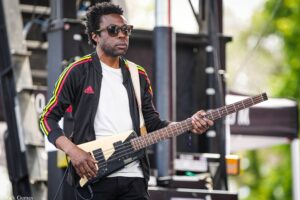 Male musician wearing sunglasses and a black jacket with red, yellow, green stripes plays a bass guitar on an outdoor stage.