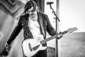 Male guitarist in a denim jacket playing an electric guitar on stage, microphone stand nearby (black-and-white photo).
