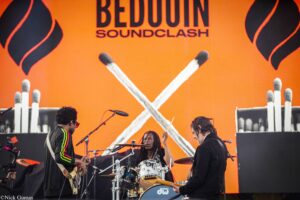 On an orange stage backdrop reading 'BEDOO N SOUNDCLASH' with three musicians playing: guitarist left, drummer center, guitarist right, microphones and instruments visible