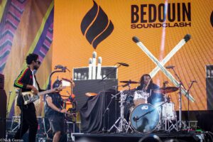 A live band performing on a stage with an orange backdrop and the text 'BEDOOIN SOUNDCLASH'; guitarist on the left, drummer at center, and other musicians with drums and equipment on stage.