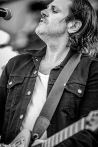 Male guitarist singing into a microphone, close-up of his face and guitar neck in black and white.