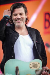 Smiling man with dark hair and stubble playing a turquoise guitar on an orange stage backdrop.