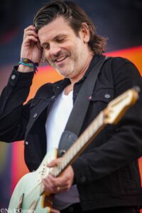 Guitarist smiling while playing a light-colored electric guitar on stage in a black jacket.