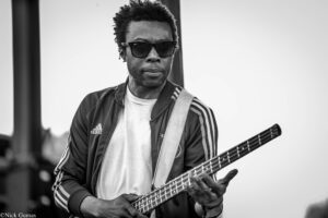 Black-and-white photo of a man wearing sunglasses and a striped jacket while playing a bass guitar on stage, looking toward the audience.