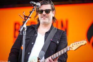 Guitarist wearing sunglasses and a black jacket singing into a microphone on an orange stage backdrop.