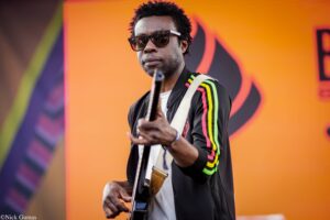 Male musician wearing sunglasses and a colorful striped jacket plays an electric guitar on a stage with an orange backdrop.
