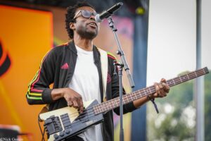 Musician wearing sunglasses and a black Adidas jacket plays a white bass guitar into a microphone on an outdoor stage, colorful background behind him.