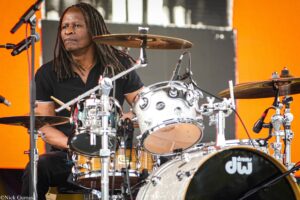 Drummer with dreadlocks playing a silver DW drum kit on stage with an orange backdrop