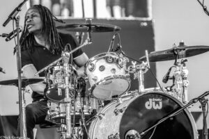 Drummer with dreadlocks performing on stage, drums and cymbals visible in a black-and-white scene.