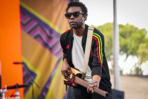 Male guitarist wearing sunglasses and a black jacket with neon stripes plays a red electric guitar in front of a colorful mural outdoors.