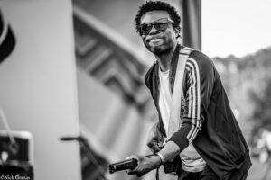 Black-and-white photo of a man wearing sunglasses and a striped jacket, playing an electric guitar outdoors with a shoulder strap in focus and the background blurred; he looks to the side and smiles slightly.