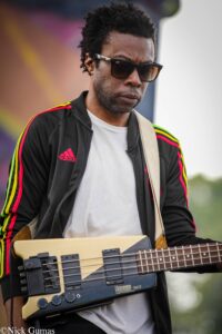 Man wearing sunglasses and a black Adidas jacket plays a yellow electric bass outdoors, gazing downward (image from Nick Gumas).