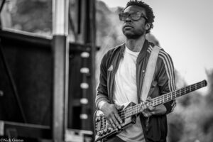 Black-and-white photo of a man wearing sunglasses and a striped jacket playing a bass guitar outdoors.