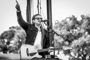 Musician performing on stage, singing into a microphone while playing a guitar; one arm raised in the air.