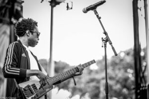 Black-and-white photo of a bass guitarist performing on stage, wearing a striped Adidas jacket on the sleeve.