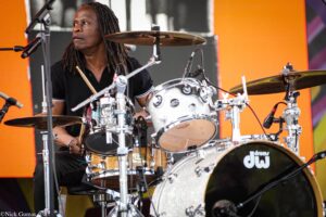 Drummer with dreadlocks playing a drum kit on stage, wearing a black polo, orange backdrop behind him, mic in front.