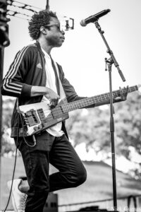 Male musician playing a bass guitar on an outdoor stage, wearing a striped Adidas jacket and sunglasses, singing into a microphone.