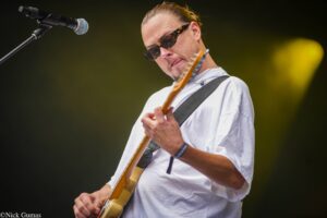 Male guitarist wearing sunglasses and a white shirt, playing an electric guitar on stage with a microphone nearby.