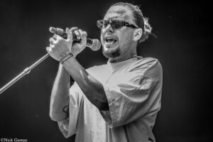 Male singer performing into a handheld microphone on stage, wearing sunglasses and a tied-back hairstyle.