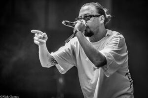 Hip-hop performer wearing sunglasses raps into a handheld microphone on stage, left arm extended toward the audience.