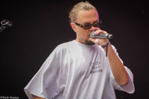 Male singer wearing sunglasses, holding a microphone on stage against a dark background.