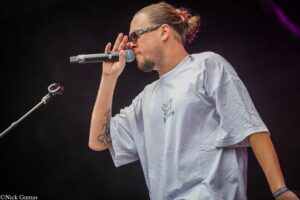 Male vocalist singing into a handheld mic on stage, wearing sunglasses and a light gray T-shirt, hair in a bun, visible tattoos on forearm.