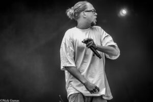 Male performer with glasses and a man bun, holding a microphone on stage amid smoke. In black and white.