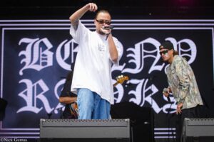 Male rapper in a white oversized shirt and jeans performs on stage, mic in hand, tattooed arm raised, Gothic backdrop behind him.