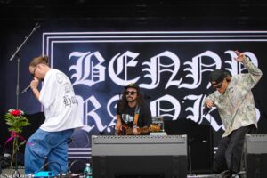 Band performing on stage: singer in white shirt on the left, guitarist center, and vocalist in camouflage jacket on the right with a large decorative backdrop behind them.