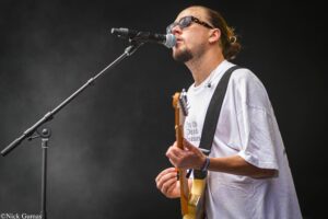 Male guitarist wearing sunglasses sings into a microphone while playing a brown electric guitar on stage in front of a dark backdrop.