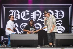 Band performing on stage with a singer in a camouflage shirt and cap, microphone in hand, raised to perform; large decorative backdrop behind them.