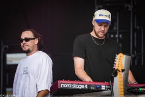 Two musicians on stage during a concert: a keyboardist in a black shirt and a cap beside a red Nord Stage 4 keyboard, with a guitarist nearby.