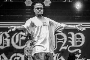 Male performer on stage with arms outstretched, wearing sunglasses; shirt reads 'On the Dead Homies' in front of a large graphic backdrop (black and white).