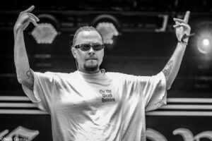 Black-and-white photo of a DJ or performer on stage, arms raised, wearing sunglasses and a light T-shirt with the text 'On the Dead Homies' visible on the chest, tattoos on the arms.
