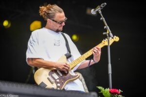 Male guitarist with a bun and sunglasses playing a light-colored electric guitar on stage, microphone stand nearby.