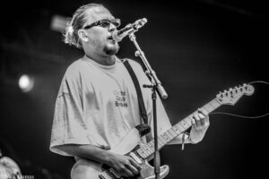 Male guitarist singing into a microphone on stage, wearing sunglasses and a light T-shirt, playing a Fender Stratocaster.