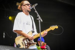 Male guitarist wearing a white t-shirt and sunglasses sings into a microphone while playing a sunburst Fender Telecaster on stage with a black strap.