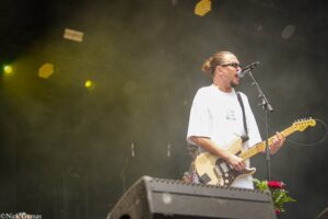 Male guitarist singing into a microphone on stage while playing a light-colored Fender Telecaster, wearing a white shirt and sunglasses amid yellow stage lights.