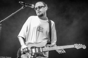 Guitarist wearing sunglasses sings into a microphone while playing a white Fender Stratocaster on stage (black‑and‑white photo).