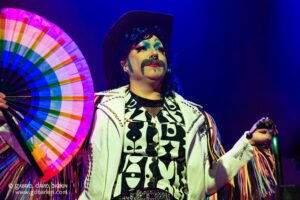 Drag performer in a white studded jacket holds a rainbow fan under blue stage lights, colorful makeup and accessories visible.