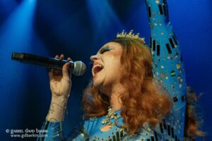 Female singer in a blue sequined outfit with a crown, singing into a handheld microphone on stage.