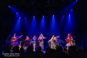 Band of colorful-clad performers with guitars and keyboards performing on a dark stage under blue spotlights.