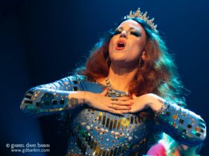 Drag queen wearing a jeweled crown and blue sequin gown, singing with hands on her chest on stage.