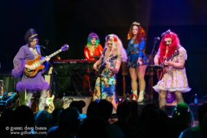 Drag performers on a lit stage playing guitars during a live show, in colorful flamboyant outfits and wigs.
