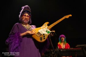 Drag performer in a purple gown and red glasses holds an electric guitar on a dark stage, with a keyboard player visible in the background.