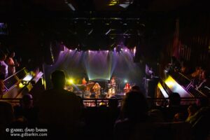 Concert stage with a band performing under colorful purple, yellow, and white lights; audience along balconies on either side in a dim venue. Watermark text visible at bottom left.