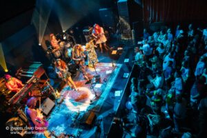 Overhead view of a lively band performing on stage with colorful outfits while a standing audience watches from the right side of the venue.",