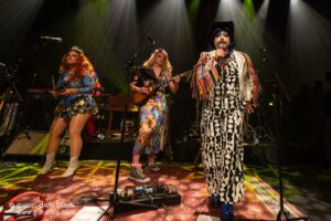 Three drag performers on stage: a blue-dressed vocalist, a floral-print guitarist, and a bold black-and-white patterned-suit singer at the microphone under stage lights.