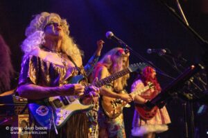 Live concert scene with a woman in a shiny purple dress and blonde wig playing an electric guitar at the front, others perform on stage under colorful lights.